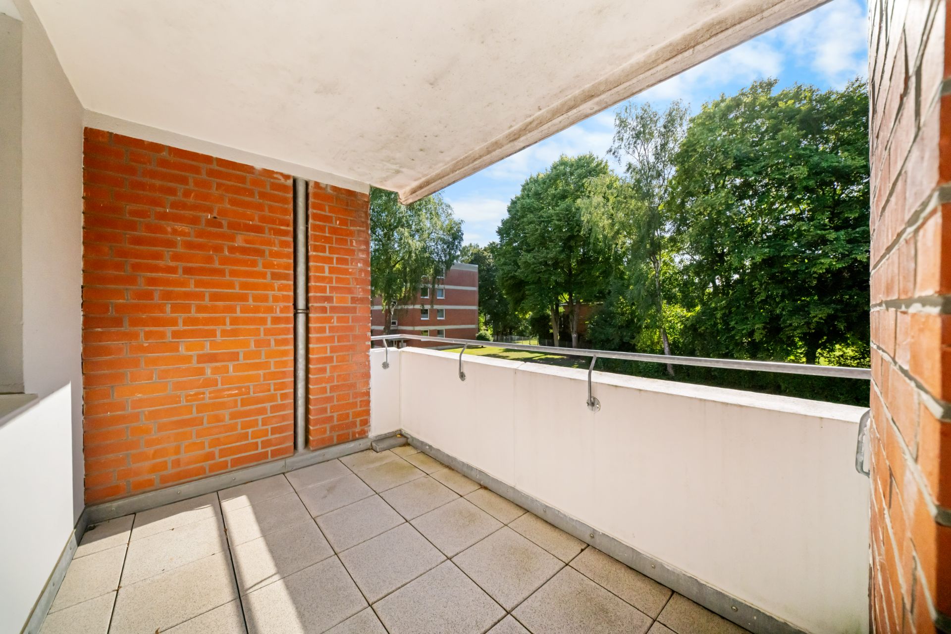Überdachter Balkon mit Fliesenboden und Blick auf Bäume und ein Wohnhaus.