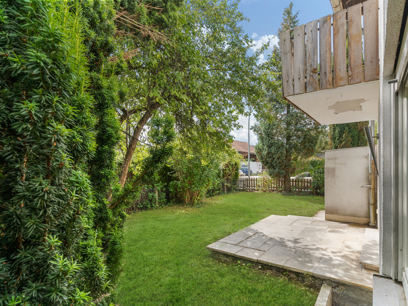 Garten mit Rasenfläche, Terrasse und Balkon, umgeben von Bäumen und Sträuchern.