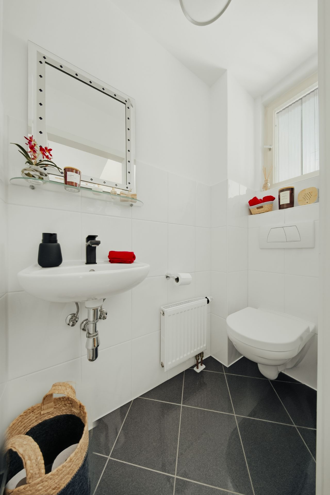 Kleines, weiß gefliestes Bad mit Wandtoilette, Waschbecken, Spiegel und Fenster.