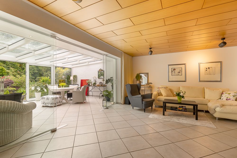 Helles Wohnzimmer mit Glaswand, beiger Couch, Sessel und Zugang zu überdachter Terrasse mit Gartensicht.