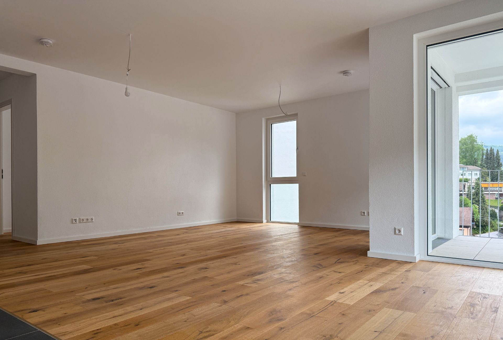 Leeres Wohnzimmer mit Holzboden, weißen Wänden und großem Fenster zur Terrasse.