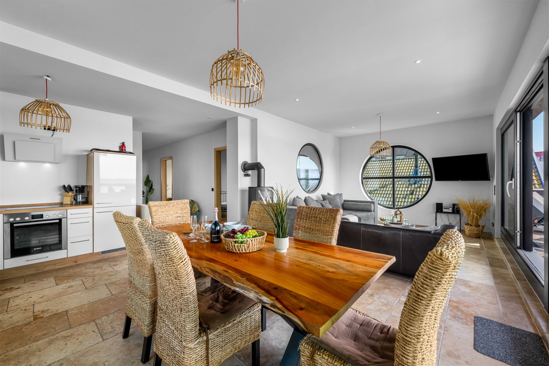 Helles Wohn- und Esszimmer mit Holztisch, Korbstühlen, moderner Küche und großen runden Fenstern.