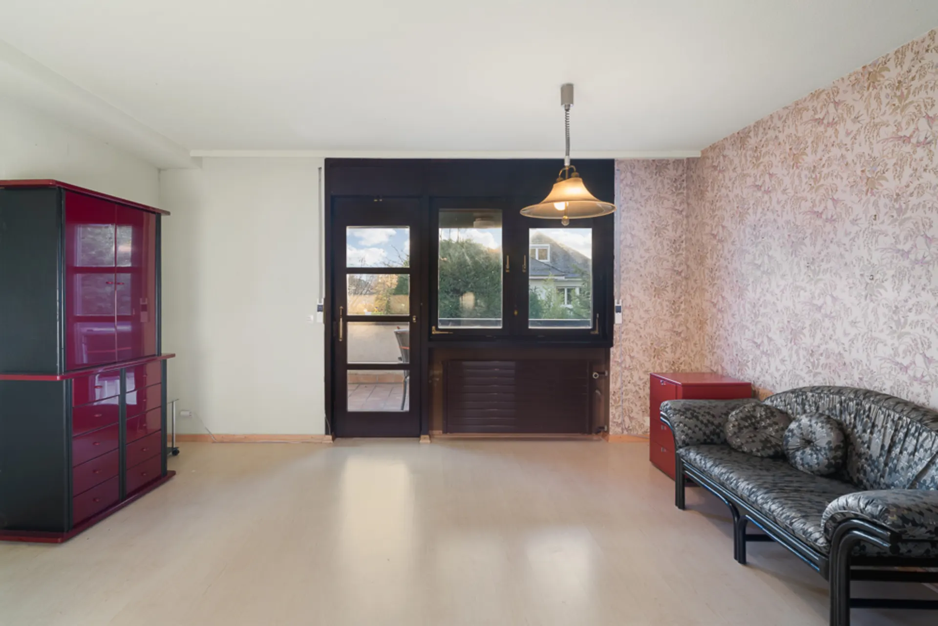 Wohnzimmer mit Sofa, Hängelampe, Schrank und Zugang zum Balkon, Tapeten an der rechten Wand.