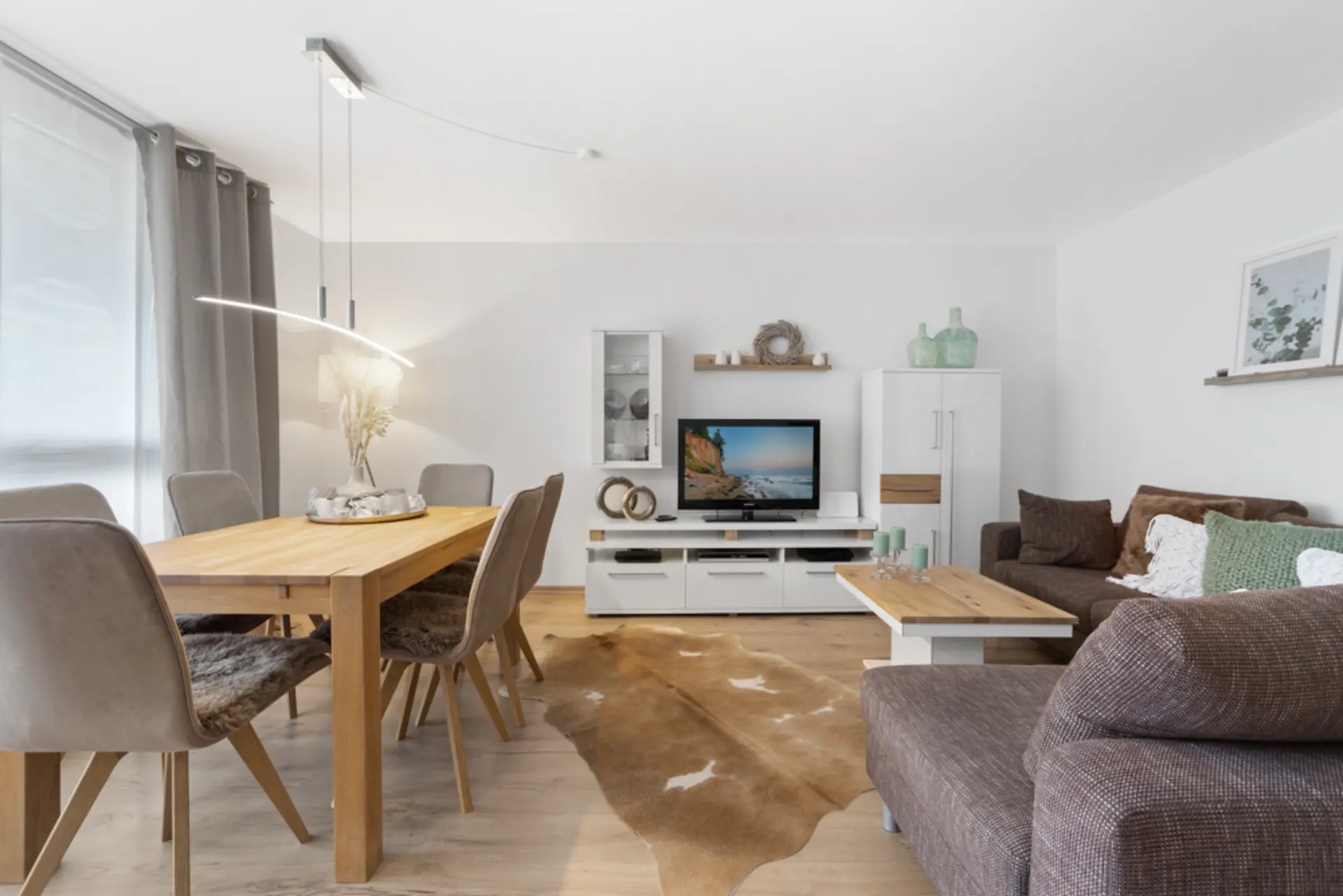 Helles Wohnzimmer mit Essbereich, Holztisch, braunen Sofas und TV auf weißem Möbel.
