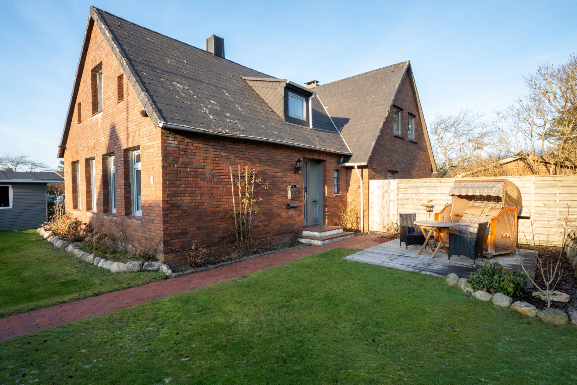 Backsteinhaus mit Garten, Terrasse und Strandkorb, umgeben von Rasen und Holzzaun.