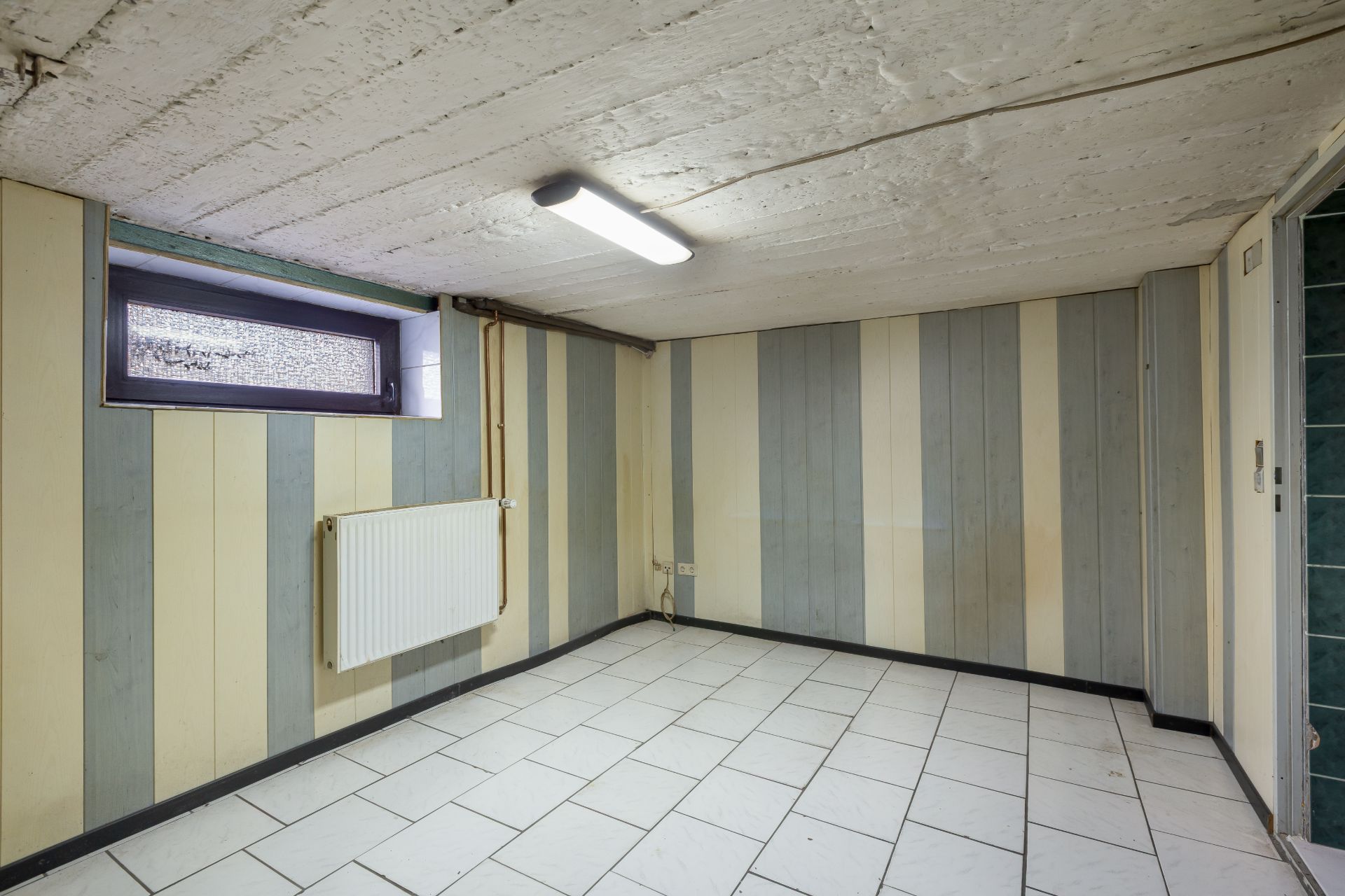 Kleiner Raum mit gestreiften Wänden, Fliesenboden, Heizkörper und kleinem Fenster.