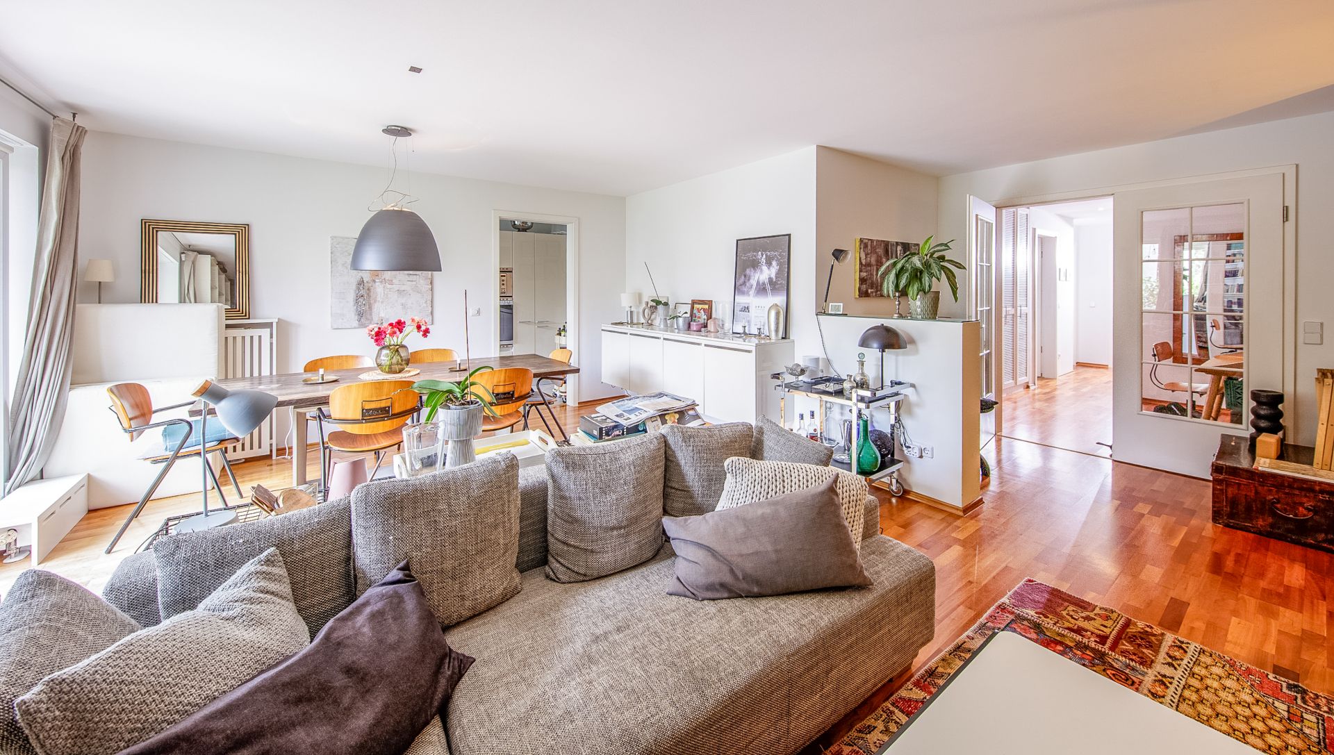 Helles Wohnzimmer mit grauem Sofa, Esstisch, Holzfußboden und großen Fenstern.