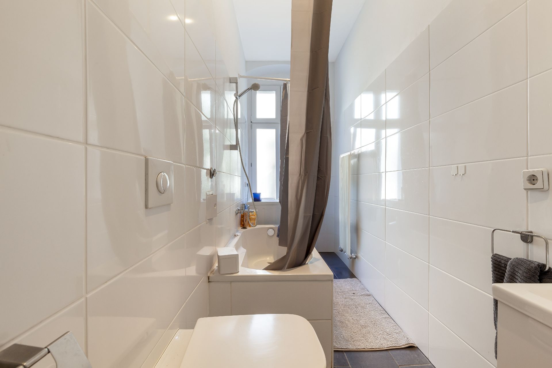 Schmales Badezimmer mit Badewanne, Duschvorhang, Fenster und weißen Fliesen.