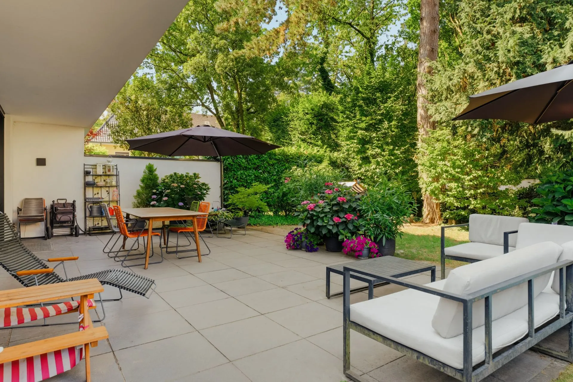 Große Terrasse mit Sitzgelegenheiten, Tischen, Sonnenschirmen und Pflanzen, umgeben von Garten und Bäumen.