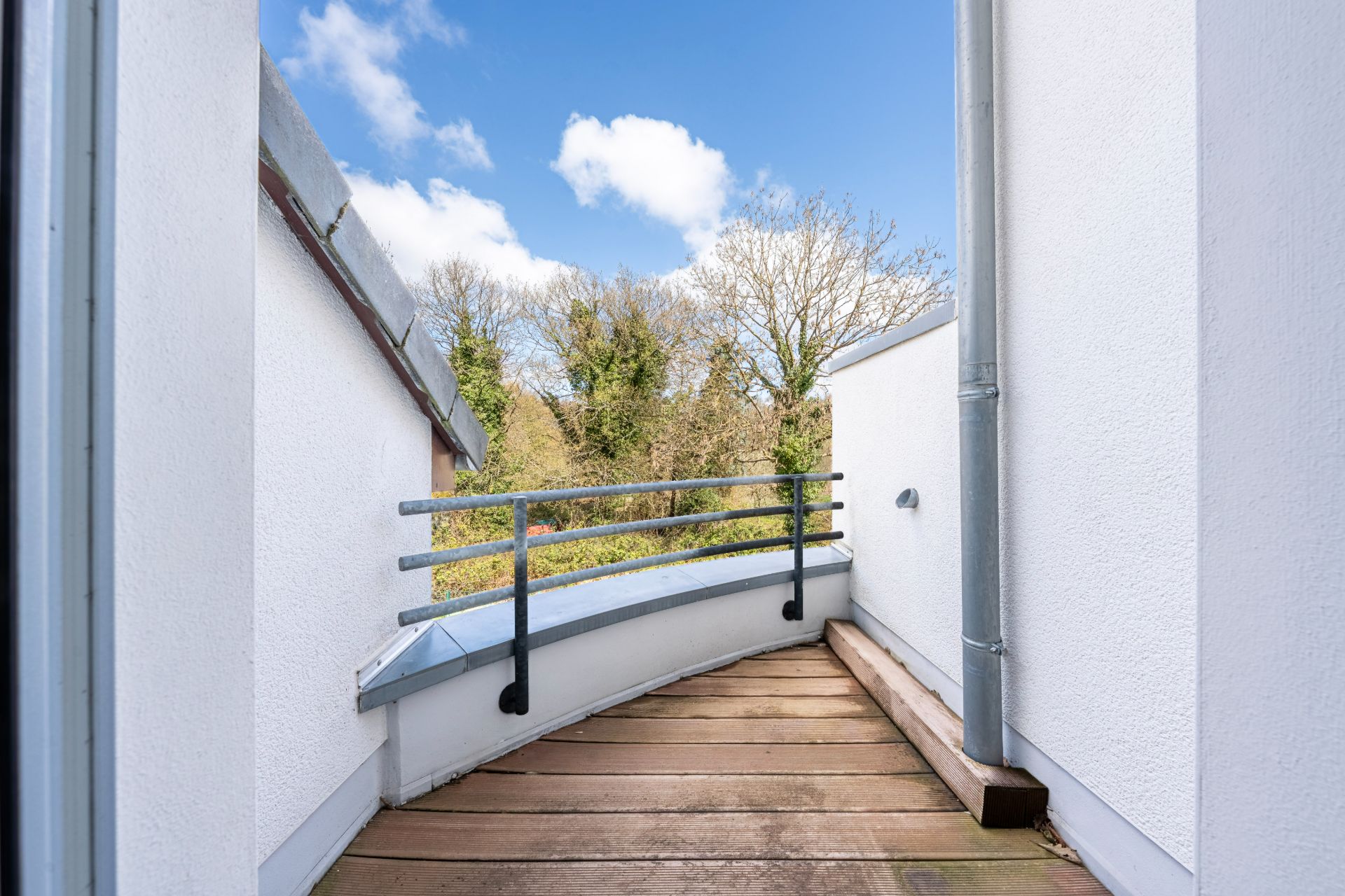 Kleine Holzterrasse mit Metallgeländer zwischen weißen Wänden, Blick auf Bäume und blauen Himmel.