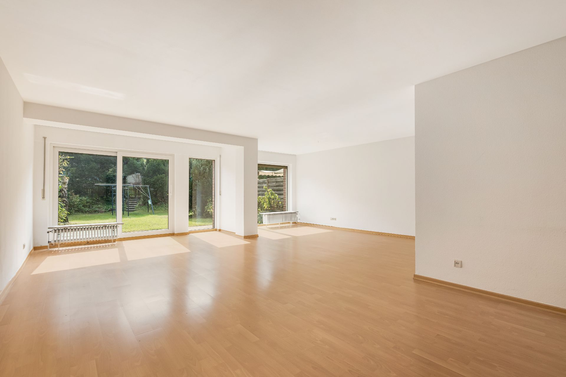 Helles, leeres Wohnzimmer mit großen Fenstern und Blick in einen grünen Garten.