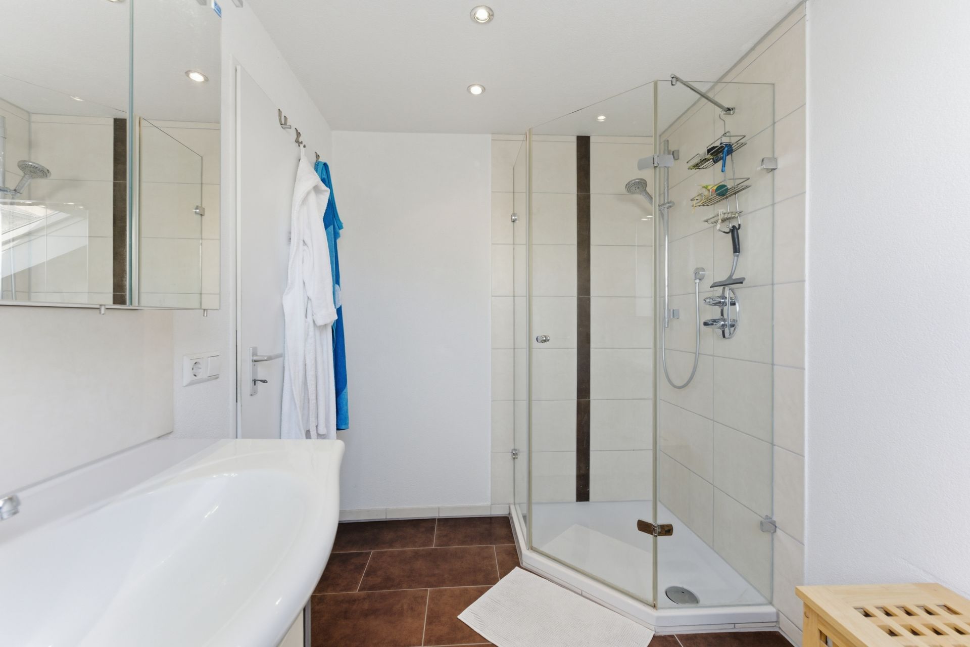Modernes Badezimmer mit Glasdusche, Badewanne, braunen Fliesen und zwei Bademänteln an der Wand.