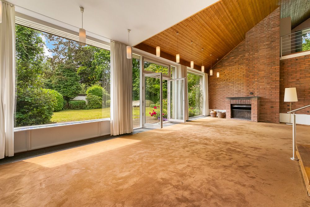Großes Wohnzimmer mit braunem Teppich, Kamin, großen Fenstern und Blick in den Garten.