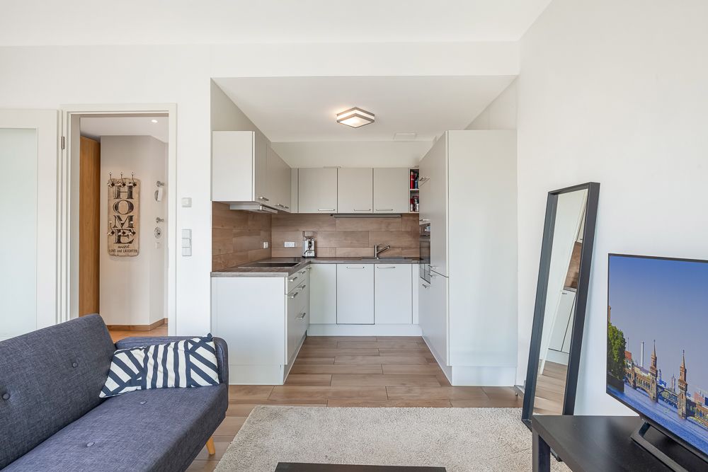 Offene, helle Küche mit weißen Schränken, angrenzend an Wohnbereich mit Sofa und Fernseher.