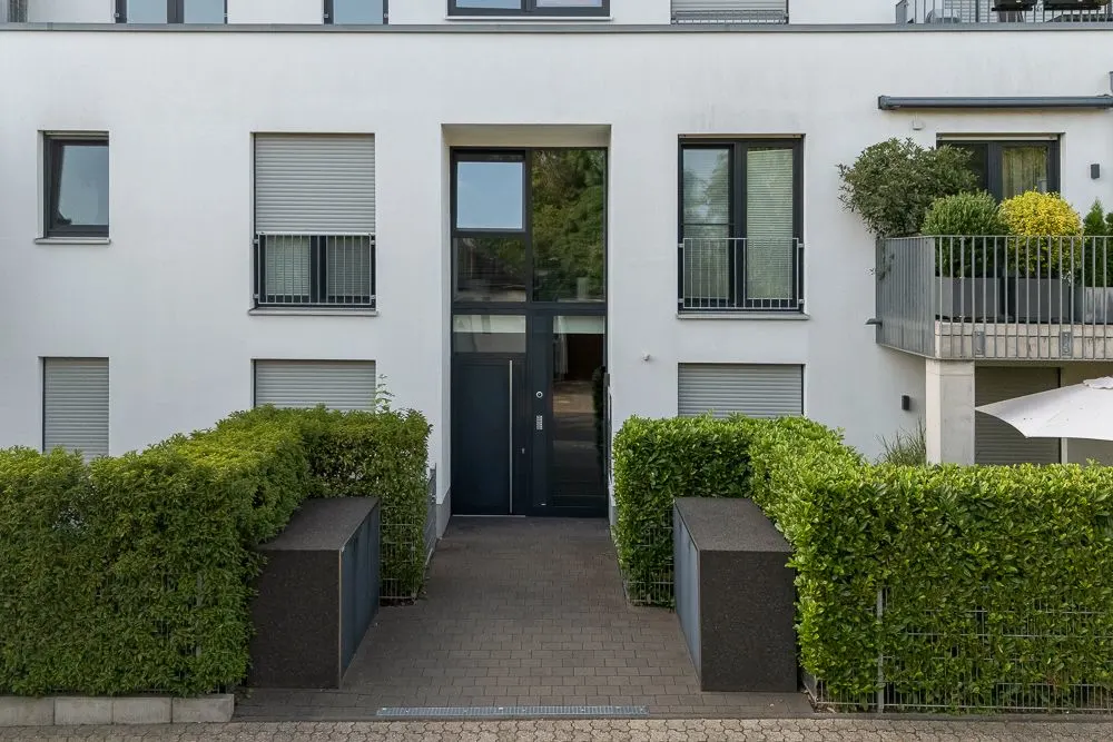 Eingangsbereich eines modernen Wohngebäudes mit Hecken und Balkon mit Pflanzen.