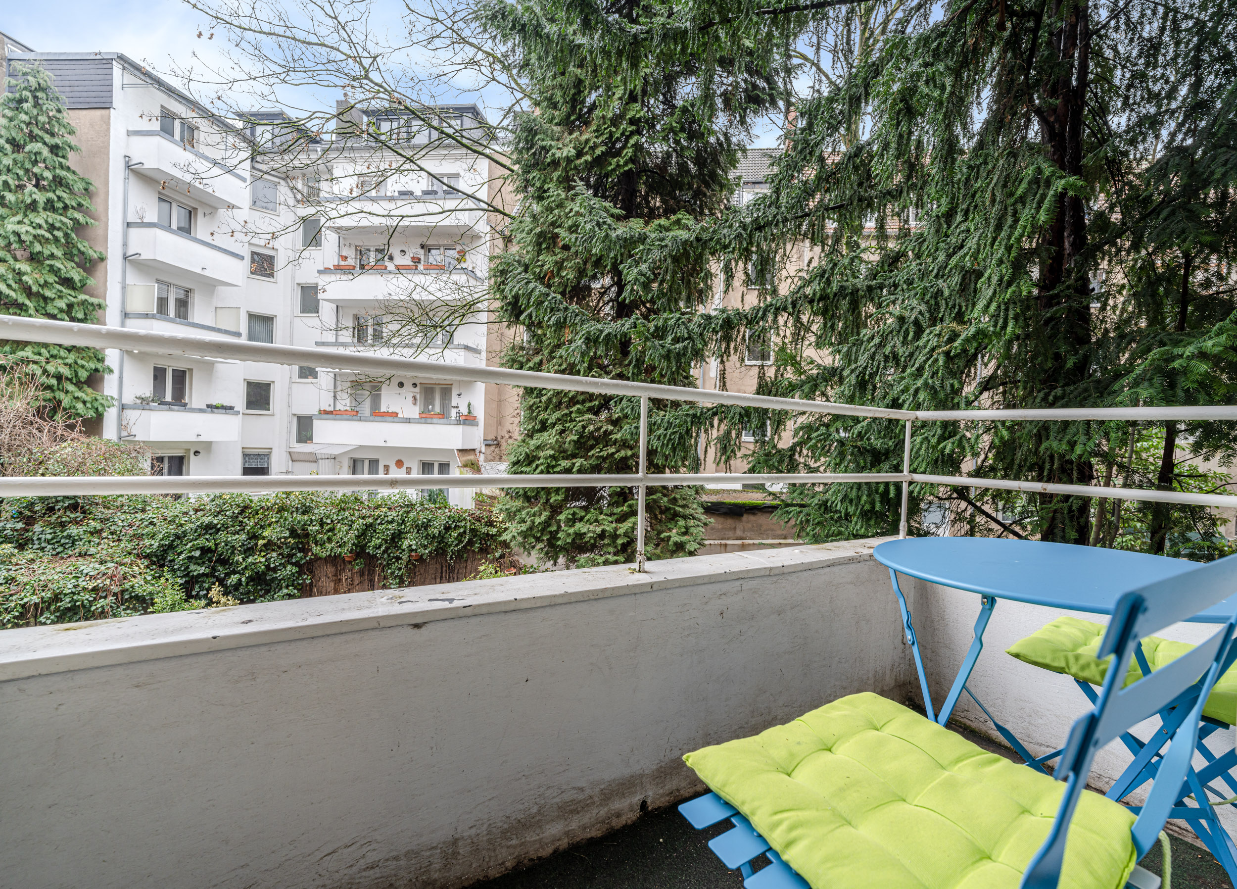 Balkon mit blauem Tisch und Stühlen, Blick auf Bäume und benachbarte Wohngebäude.