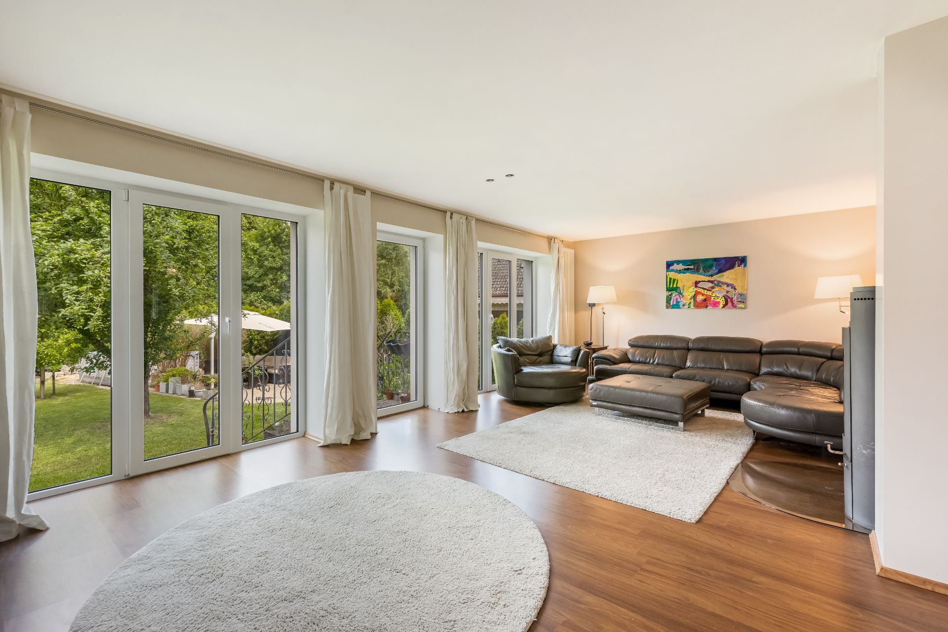 Helles Wohnzimmer mit großen Fenstern, Ledersofa, Teppichen und Blick auf den Garten.