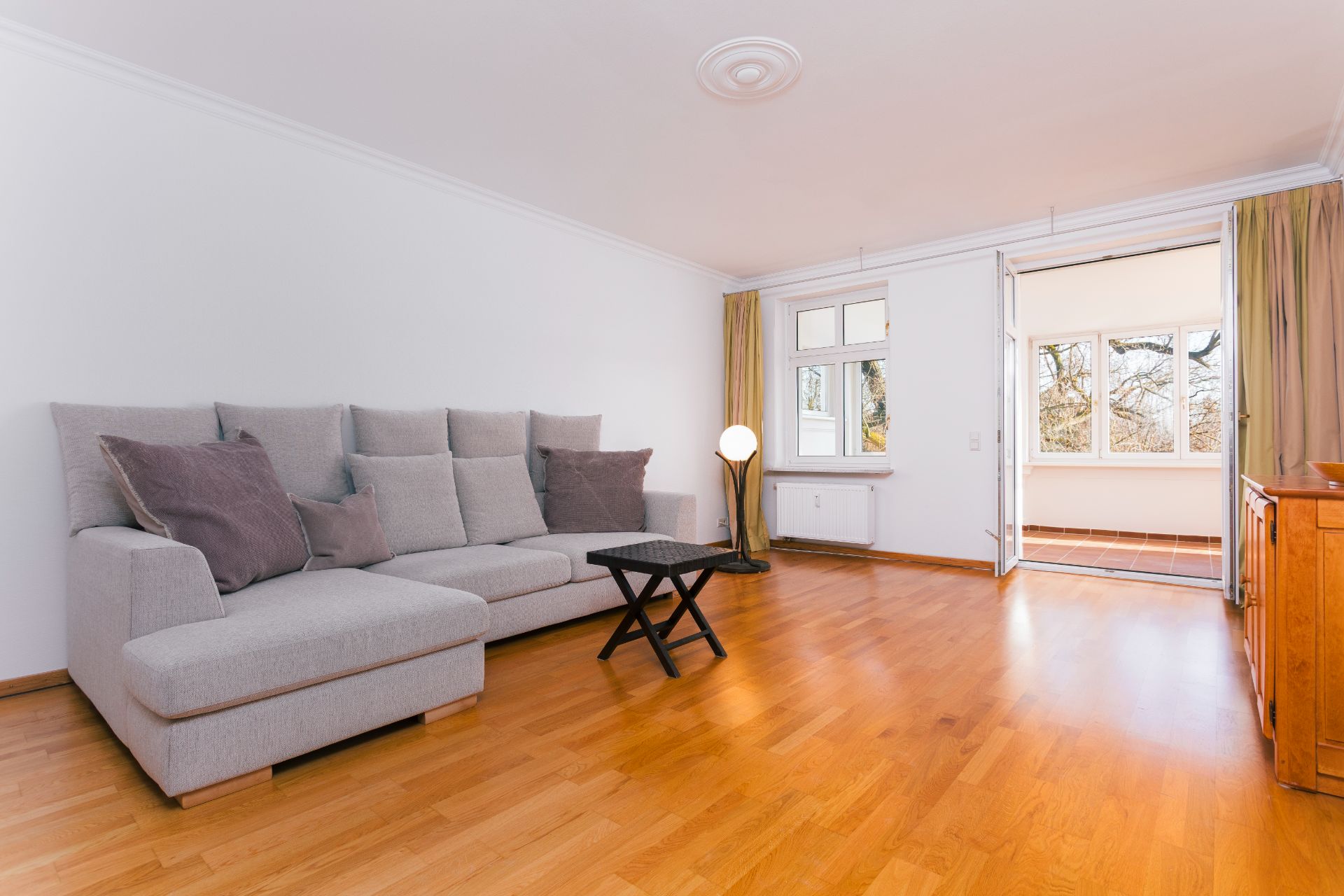 Helles Wohnzimmer mit Ecksofa, kleinem Tisch, Holzfußboden und Zugang zu einem verglasten Balkon.