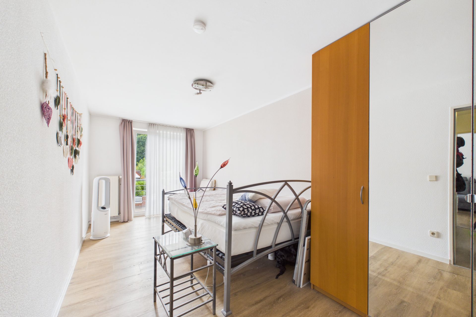 Helles Schlafzimmer mit großem Fenster, Metallbett, Nachttisch und Spiegelschrank.