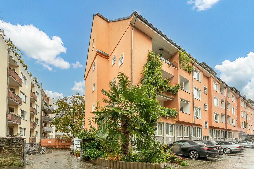 Mehrfamilienhaus mit oranger Fassade, Balkonen und grünen Pflanzen, daneben parkende Autos.
