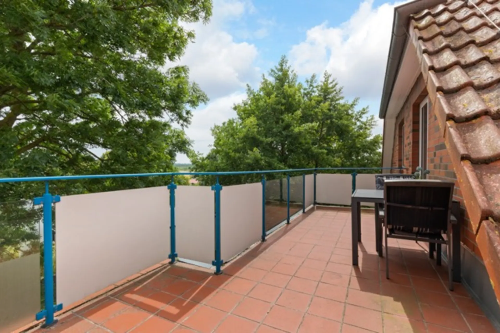Großer Balkon mit Terrakotta-Fliesen, blauem Geländer und Sitzgruppe, umgeben von Bäumen.