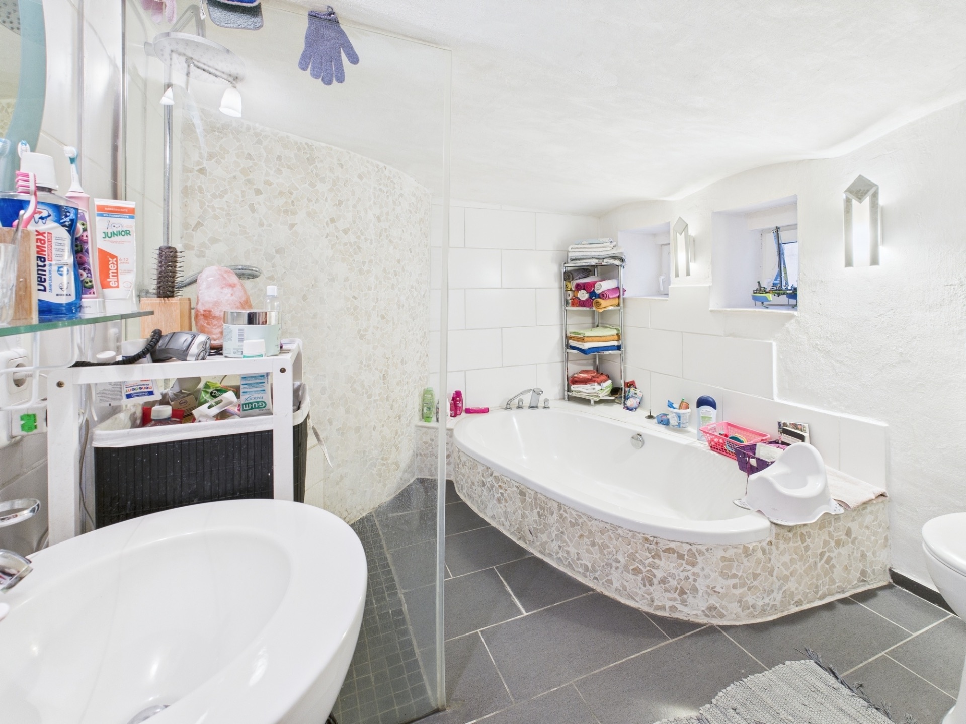 Helles Badezimmer mit Eckbadewanne, Waschbecken, Dusche und Regalen mit Handtüchern.