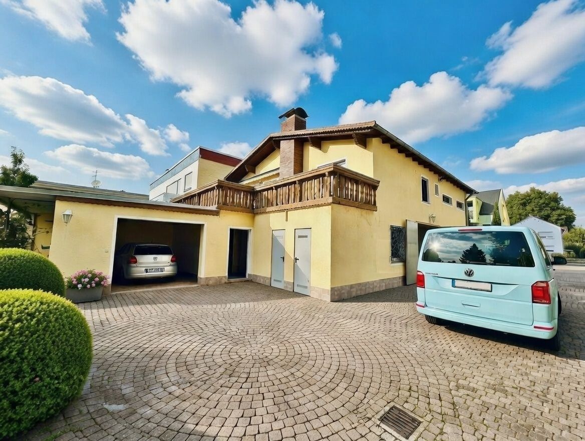 Gelbes Wohnhaus mit Garage und gepflastertem Hof, zwei Autos sind sichtbar.
