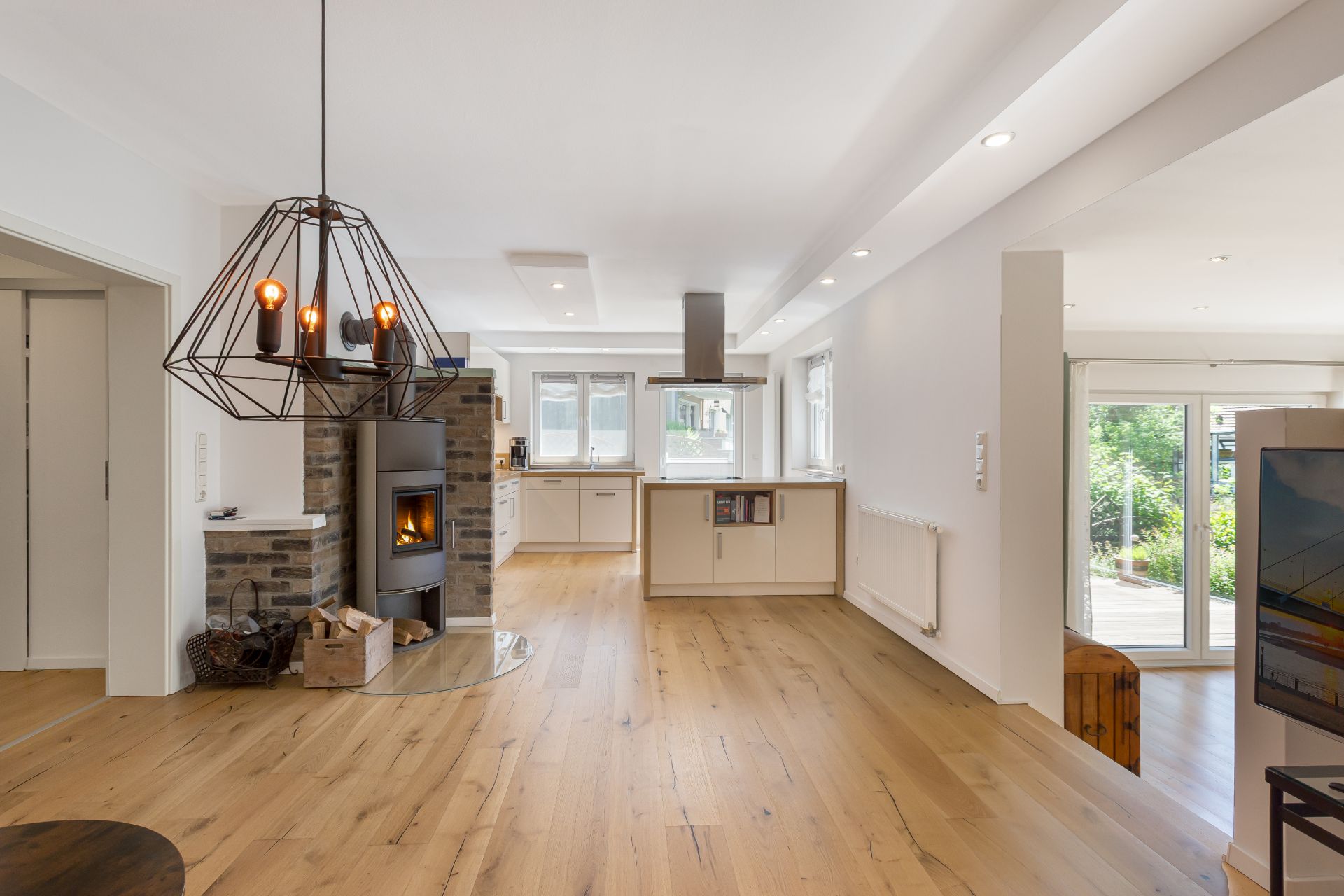 Helles Wohnzimmer mit Holzboden, Kaminofen, offener Küche und großen Fenstern.