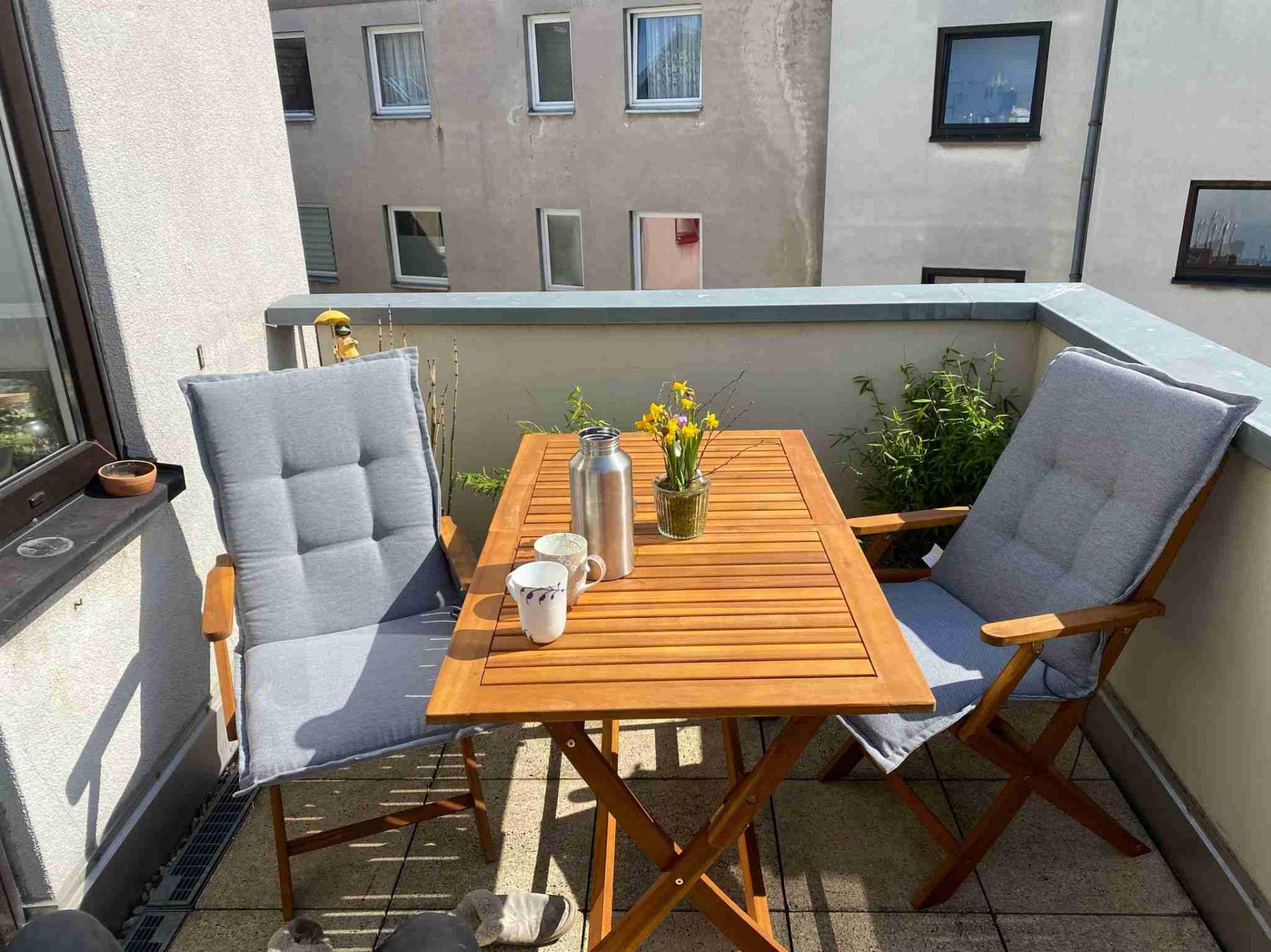 Balkon mit Holztisch, zwei Stühlen mit Polstern und Blumen, Tasse und Thermoskanne.