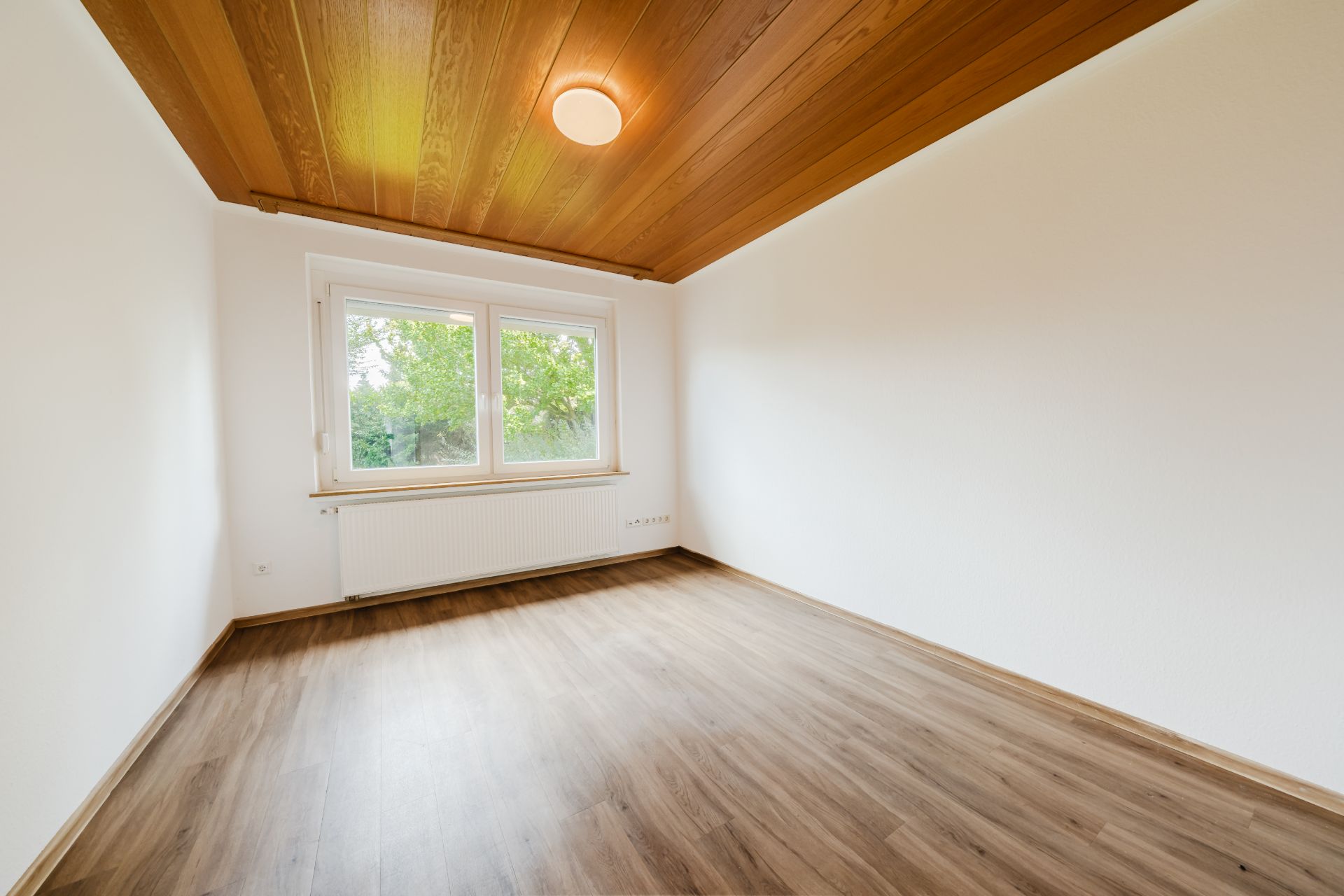 Leeres Zimmer mit Holzdecke, großem Fenster und Laminatboden.