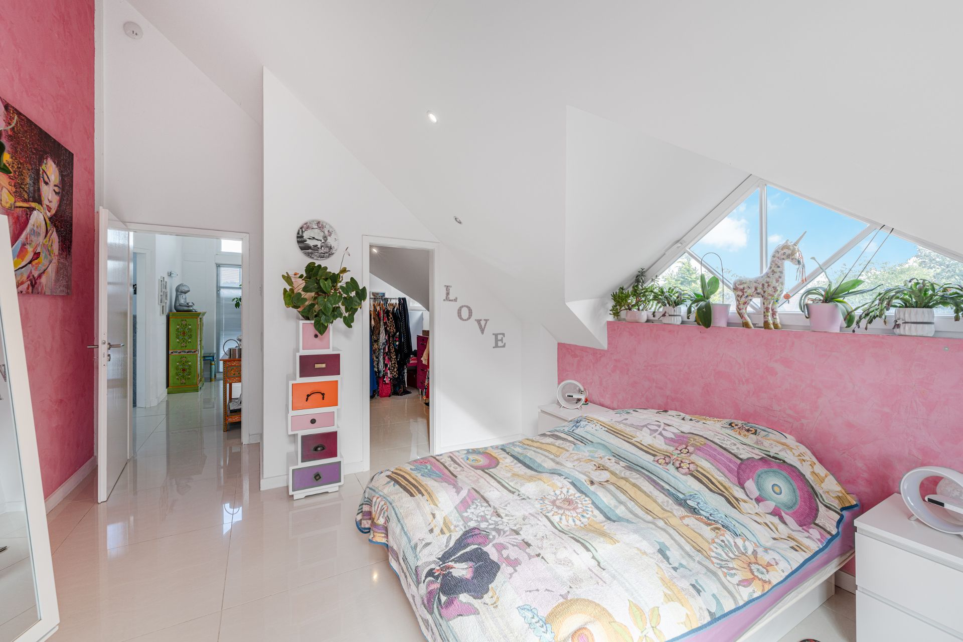 Helles Schlafzimmer mit buntem Bett, pinker Wand, Pflanzen und großem Fenster im Dachgeschoss.