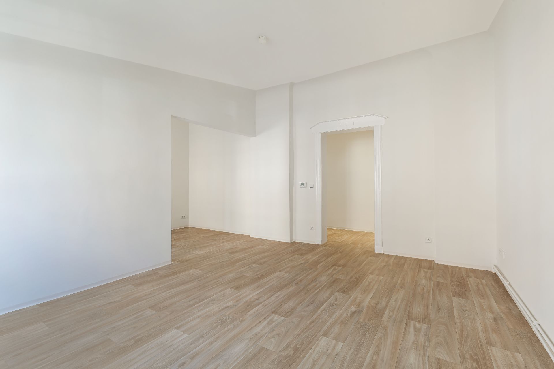 Leerer Raum mit hellem Holzboden und weißen Wänden, zwei offene Türen führen in andere Zimmer.