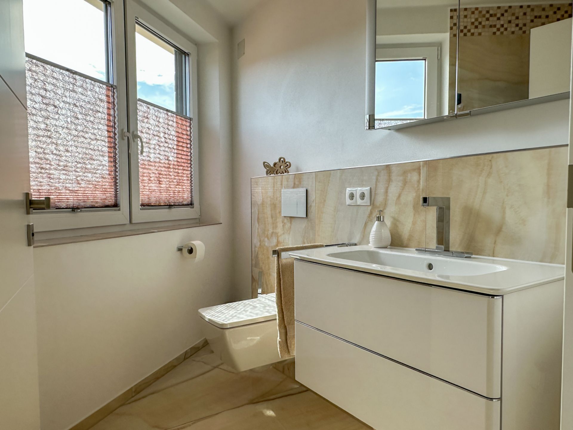 Helles Badezimmer mit Fenster, WC, Waschbecken und Spiegel.