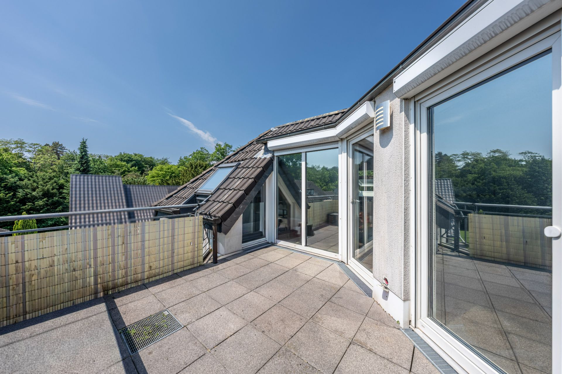 Geräumige Dachterrasse mit Fliesenboden und Glasgeländer, umgeben von Bäumen.