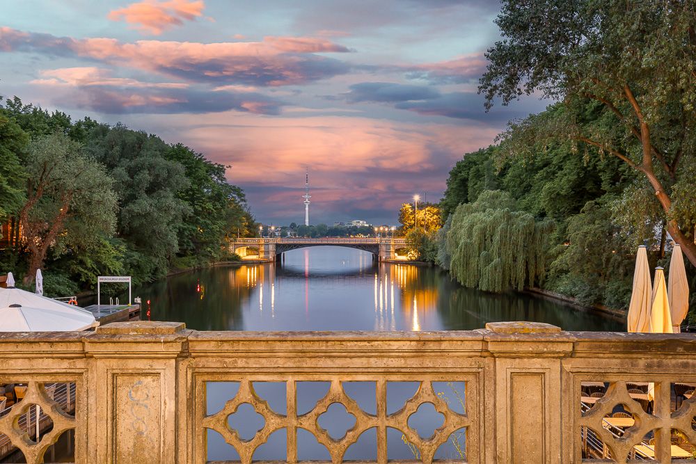 Blick von einer Steinbrücke auf einen Fluss mit Bäumen und einer Brücke im Hintergrund bei Sonnenuntergang.