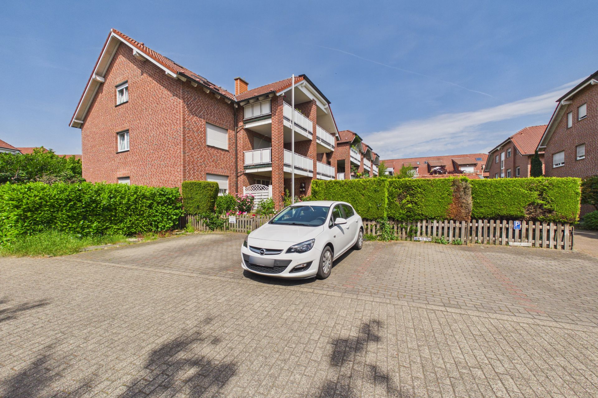 Ein weißes Auto steht auf einem Parkplatz vor einem roten Wohnhaus mit Balkon und Gartenhecke.
