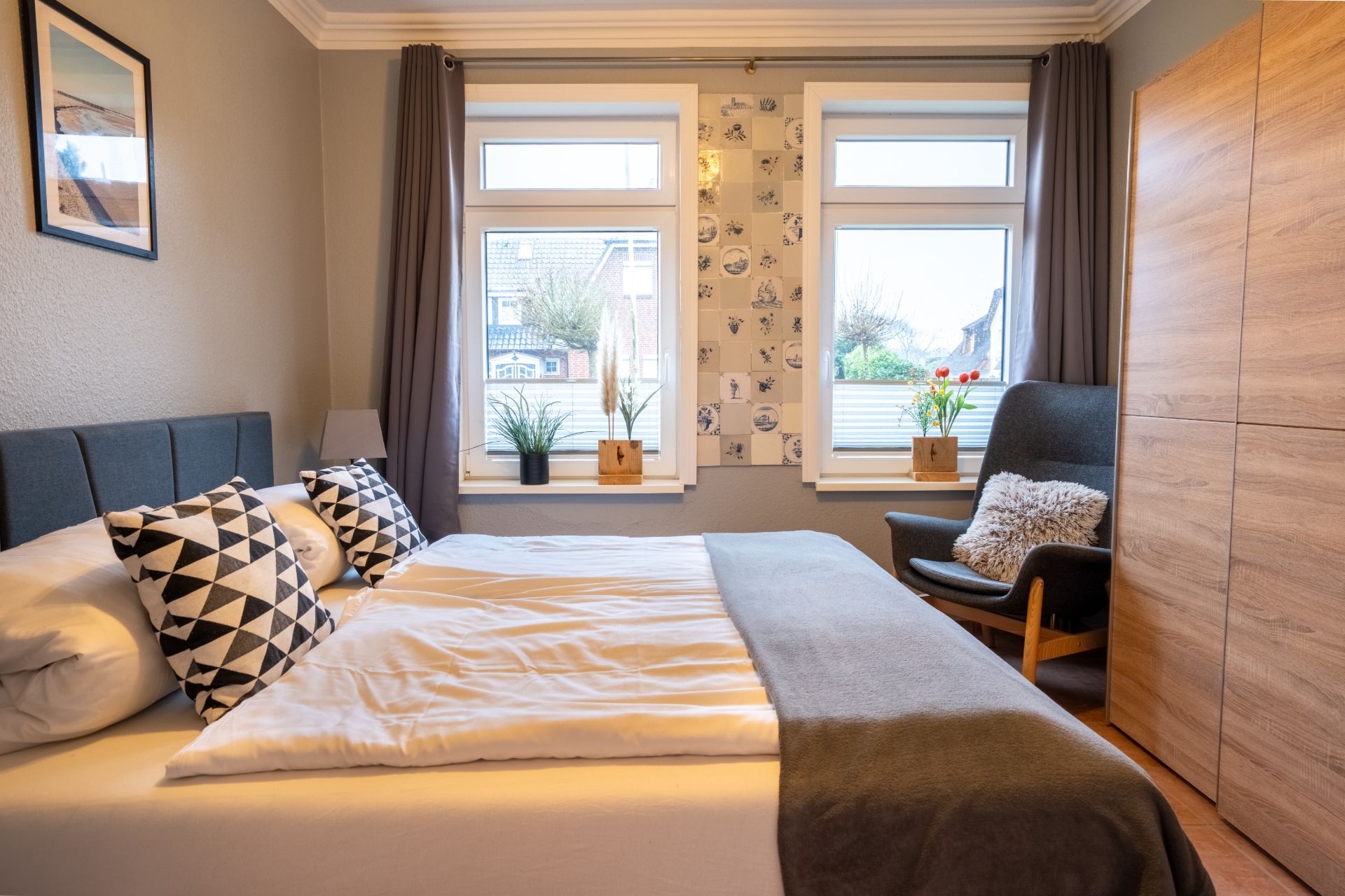 Modernes Schlafzimmer mit Doppelbett, zwei Fenstern, Sessel und großem Holzschrank.