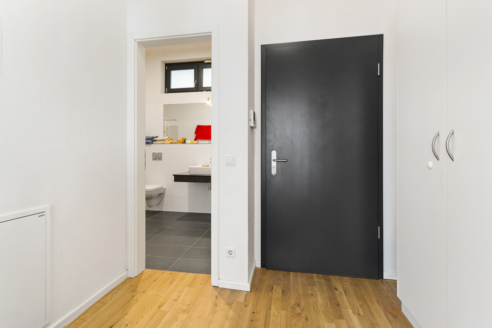 Flur mit Holzboden, schwarzer Tür und Blick ins Badezimmer mit weißen Fliesen.