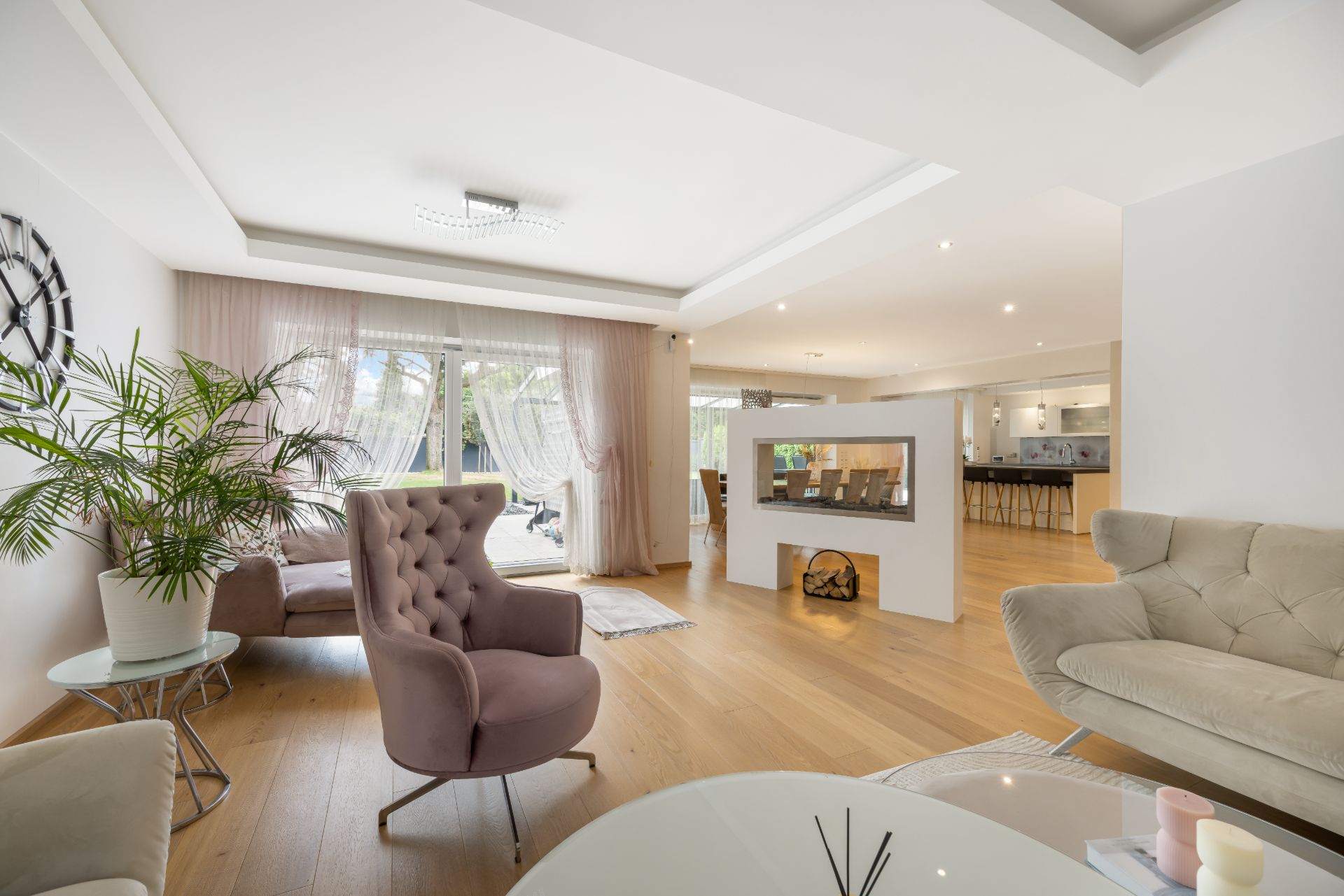 Helles Wohnzimmer mit Sesseln, Sofa, Zimmerpflanze und großem Fenster, angrenzend an Essbereich und Küche.