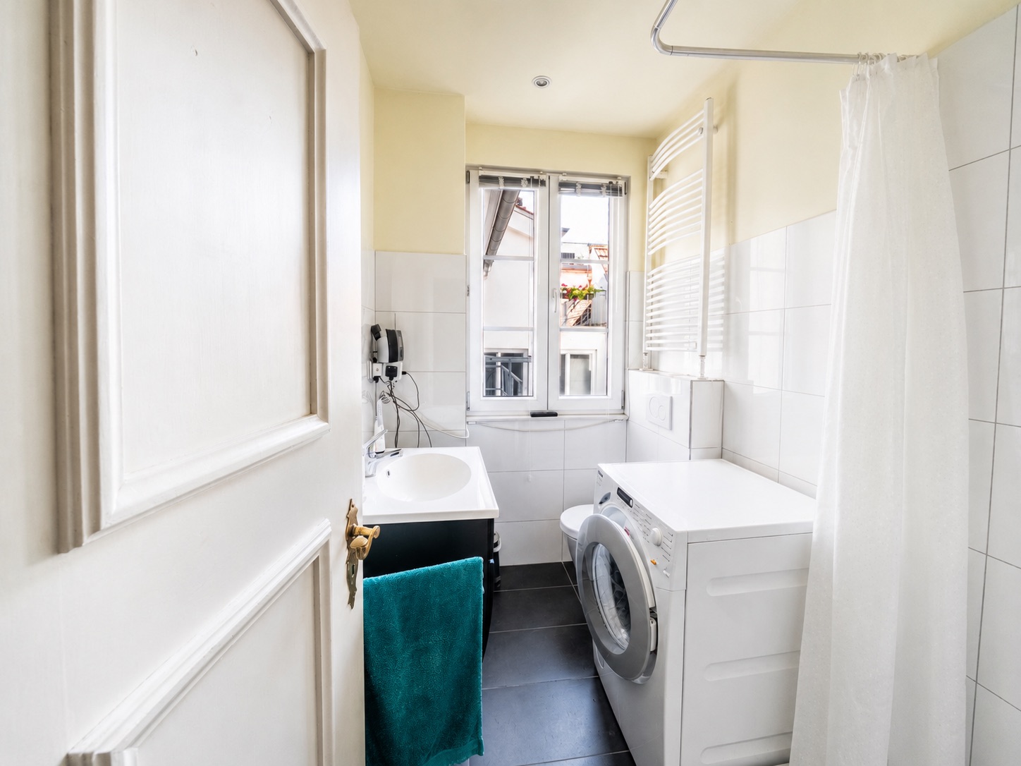 Helles Badezimmer mit Waschmaschine, Waschbecken, Dusche und Fenster.