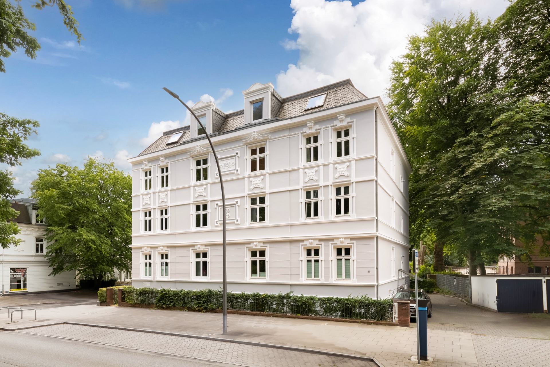 Weißes Wohngebäude mit drei Etagen und vielen Fenstern, umgeben von Bäumen.