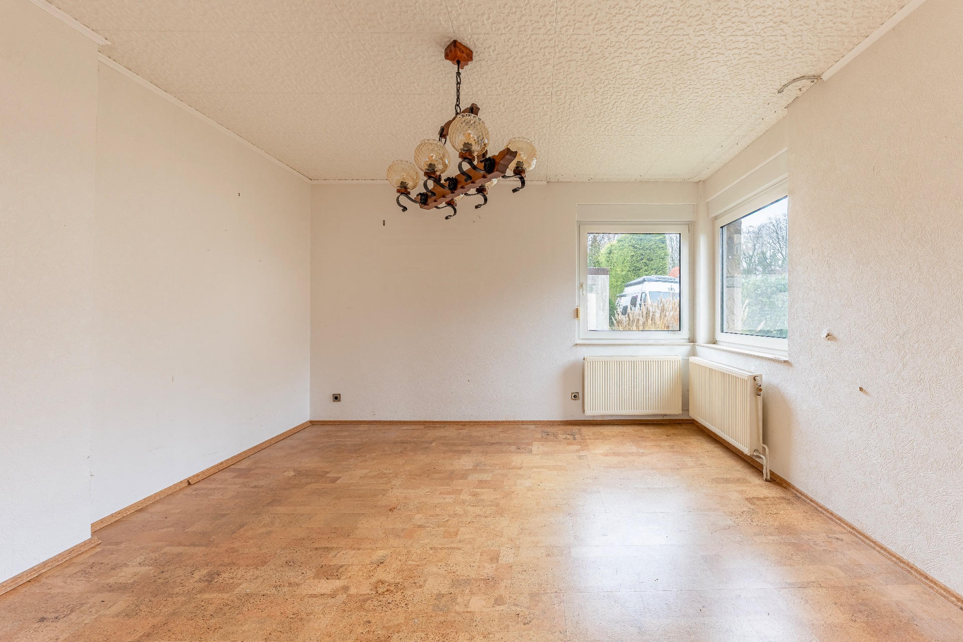 Leerer Raum mit Holzfußboden, Kronleuchter und zwei Fenstern.