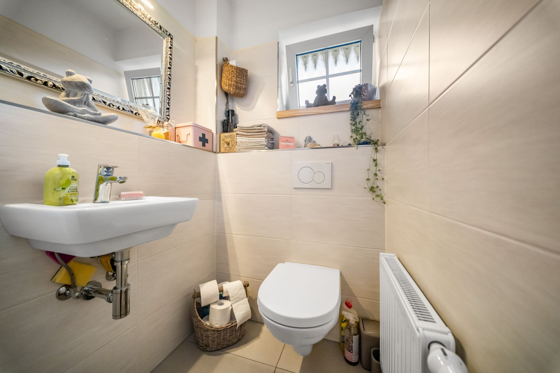 Kleines Badezimmer mit Wandtoilette, Waschbecken, Spiegel, Fenster und Heizung.