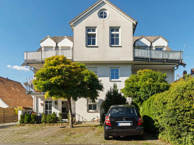 Vorderansicht eines weißen Wohnhauses mit Balkon, Bäumen und geparktem Auto vor dem Haus.