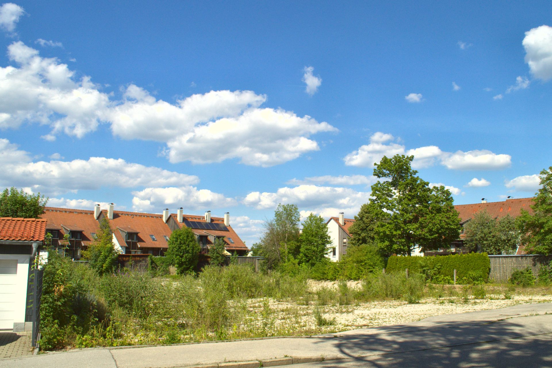 Leeres Grundstück mit Gras und Büschen, dahinter Wohnhäuser mit roten Dächern.