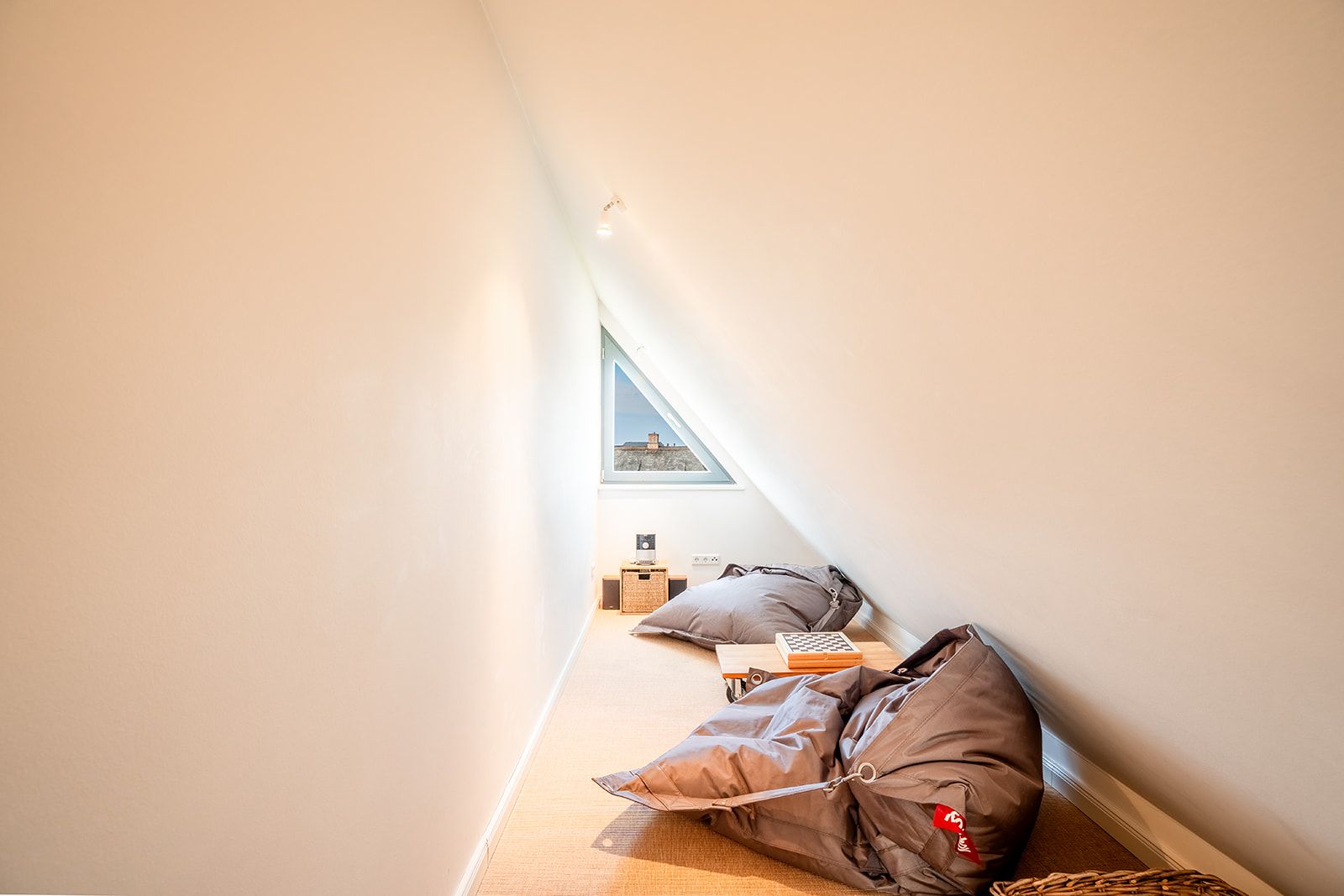 Schmaler Dachraum mit zwei Sitzsäcken, kleinem Tisch und dreieckigem Fenster.