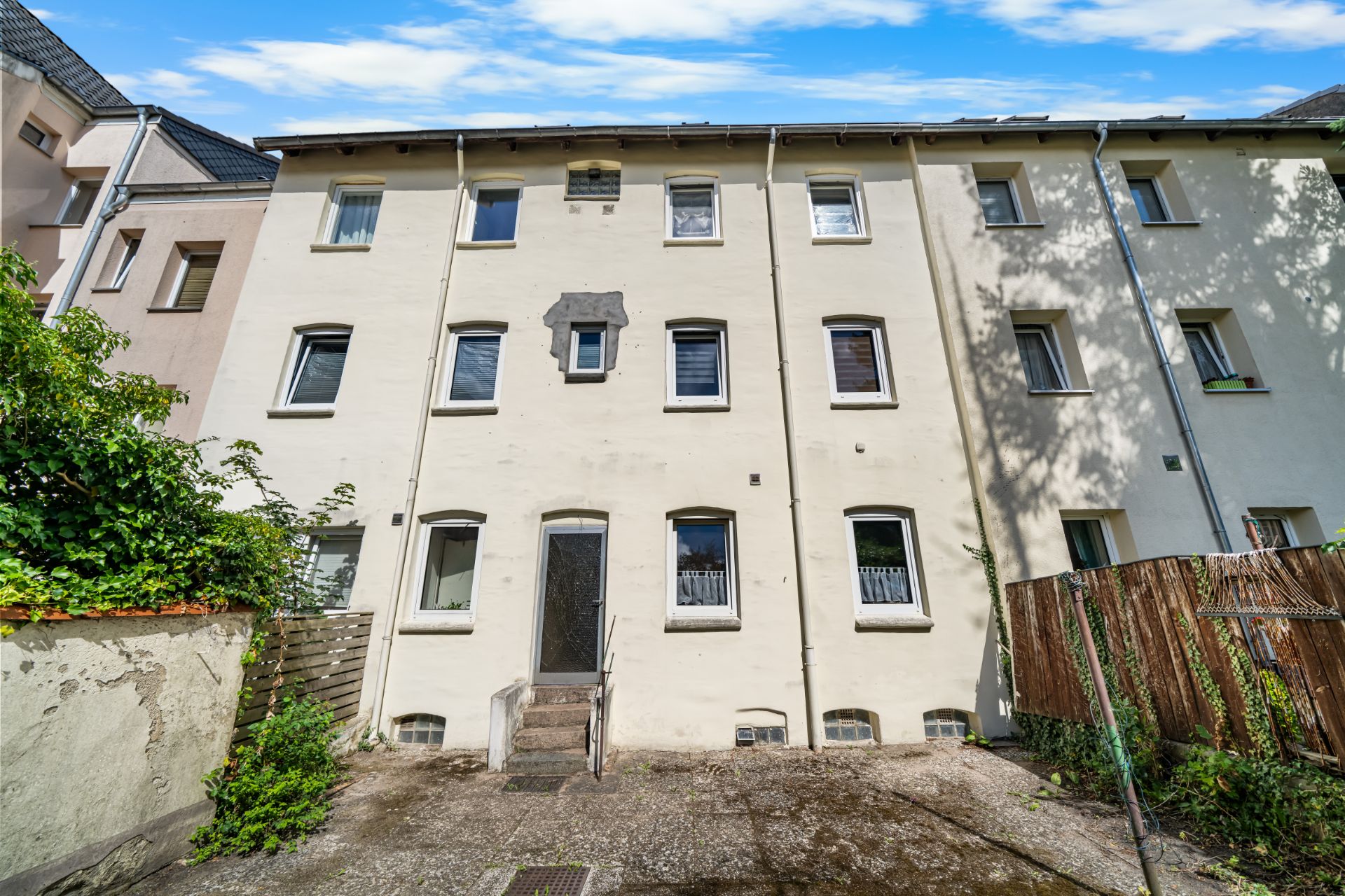 Rückseite eines hellen Mehrfamilienhauses mit mehreren Fenstern und einer Tür zum Hof.