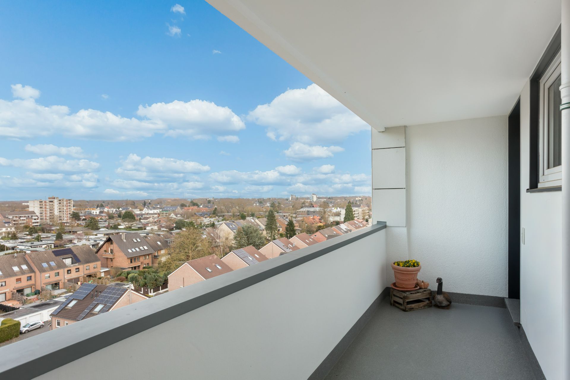 Überdachter Balkon mit Blumentopf und weiter Aussicht über Dächer und Häuser einer Wohnsiedlung.