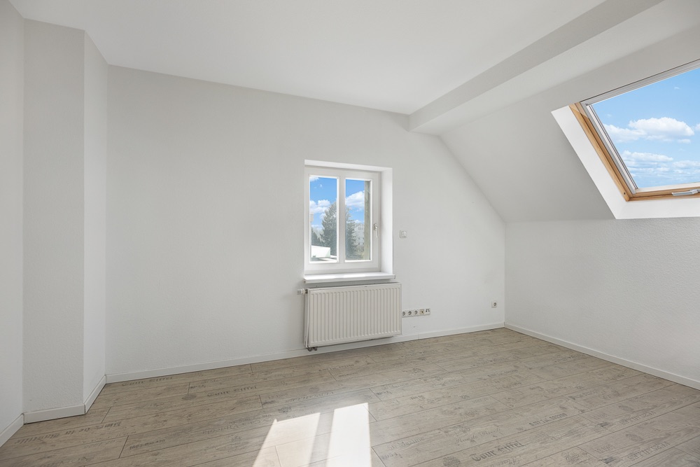 Helles Zimmer mit zwei Fenstern, hellem Holzboden und weiß gestrichenen Wänden.