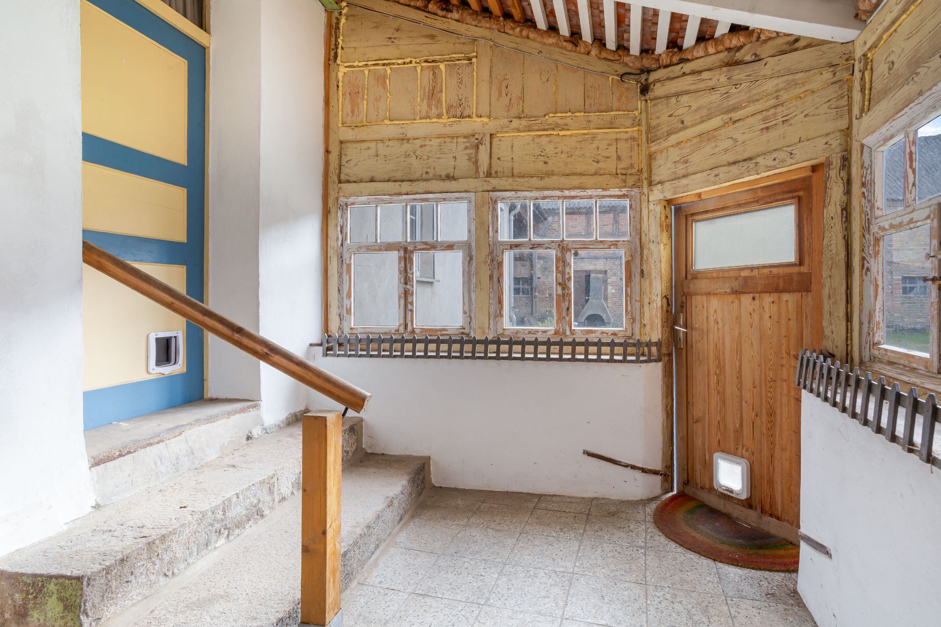 Eingangsbereich mit Holztür, Fenster, Fliesenboden und Katzenklappen an Tür und Wand.